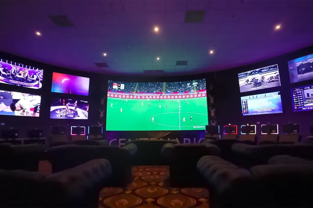 Pantallas deportivas en Casino Centenario con sillones lounge al frente.