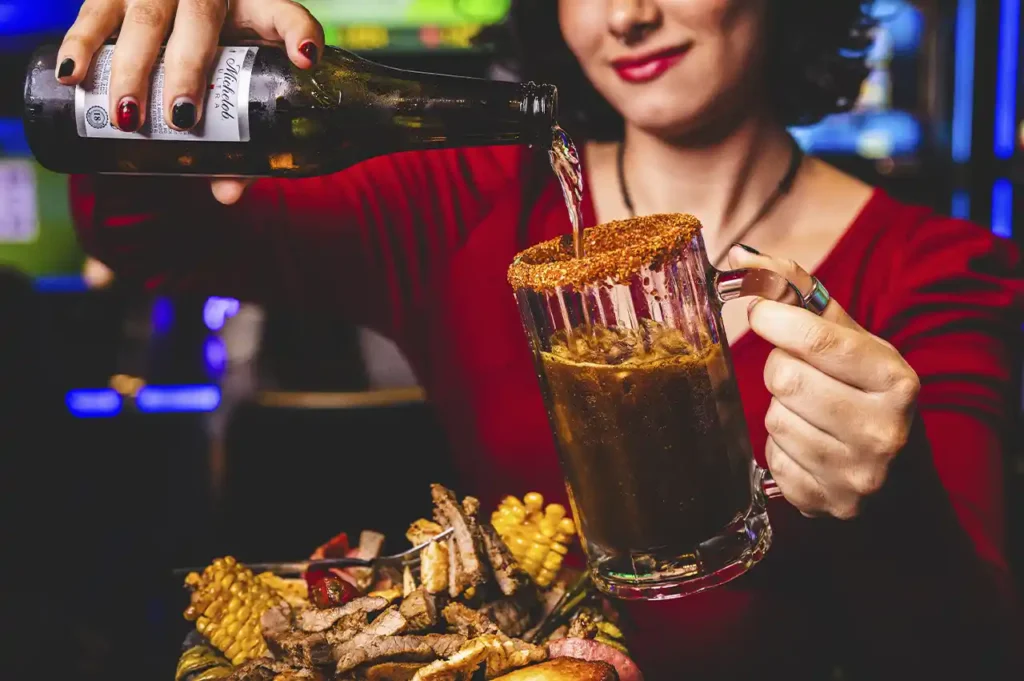 Mujer sirviendo cerveza en una michelada acompañada de botana con carne asada y elote.