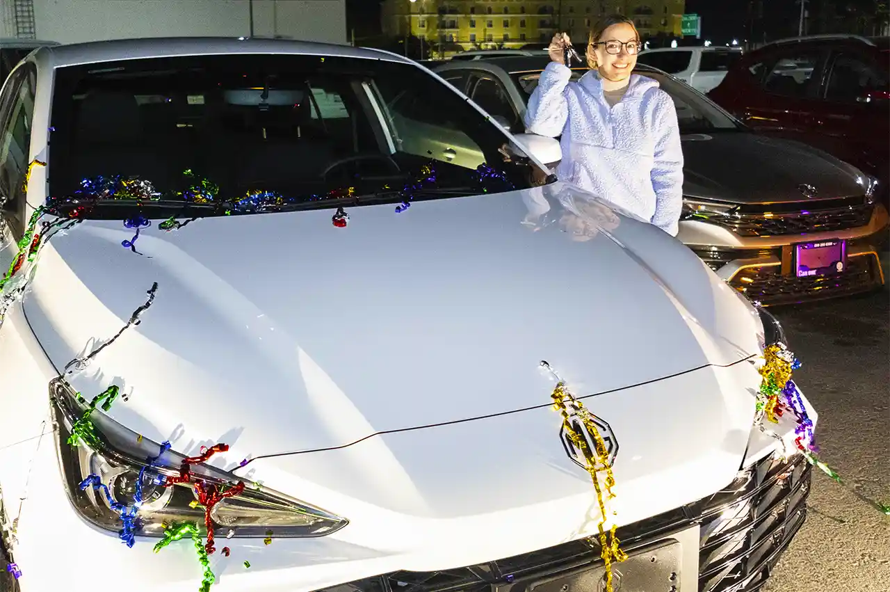 Mujer sonriente sostiene las llaves de su nuevo auto blanco decorado con serpentinas de colores.