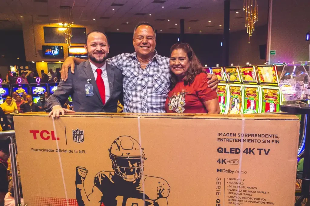 Tres personas sonrientes posan detrás de un gran televisor TCL 4K QLED, con máquinas tragamonedas iluminadas al fondo en Casino Centenario.