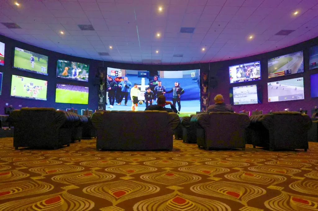 Personas viendo deportes en pantallas gigantes desde sillones lounge en Casino Centenario.